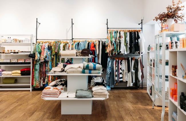 A fashion store interior with folded knitwear on a tiered shelf and racks of dresses and shirts against the wall behind