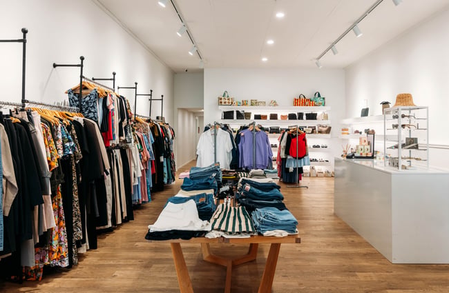 Overview of fashion store with racks of clothing against the wall and in the middle of the store. Shelves of bags and accessories are on the wall in the distance.