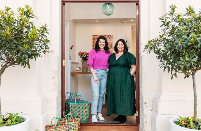 Two women standing in the doorway of Inc. Design store in Ōamaru next to colourful handwoven bags