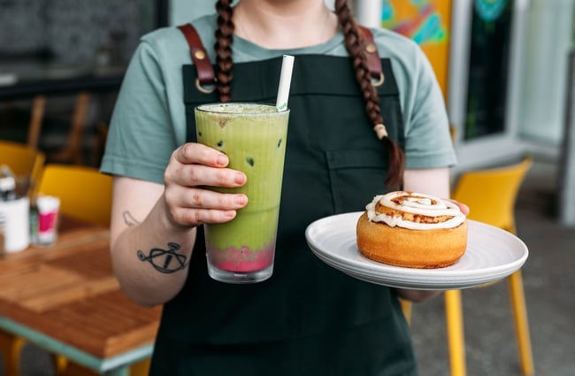 Staff member holds an iced matcha and a cinnamon roll at Kōwhai Eatery.