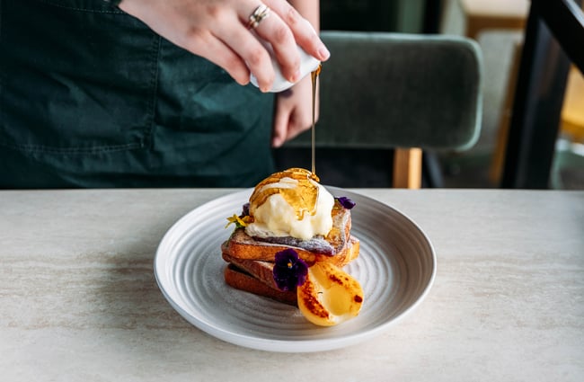 Maple syrup is being poured on a plate of French toast at Kōwhai Eatery.