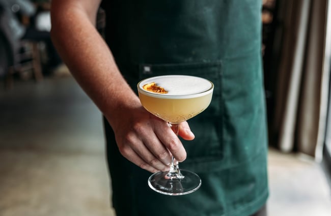 Close up of a member of staff in an apron holding out a pale yellow cocktail with a piece of dehydrated citrus placed on the white foamy top.