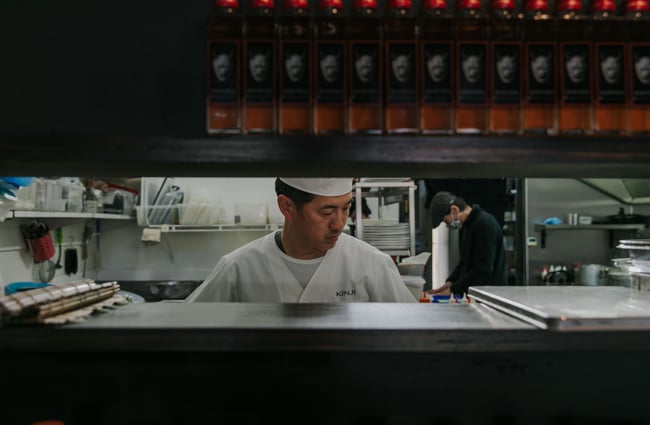 Looking over the counter into the kitchen where there are two chefs working.