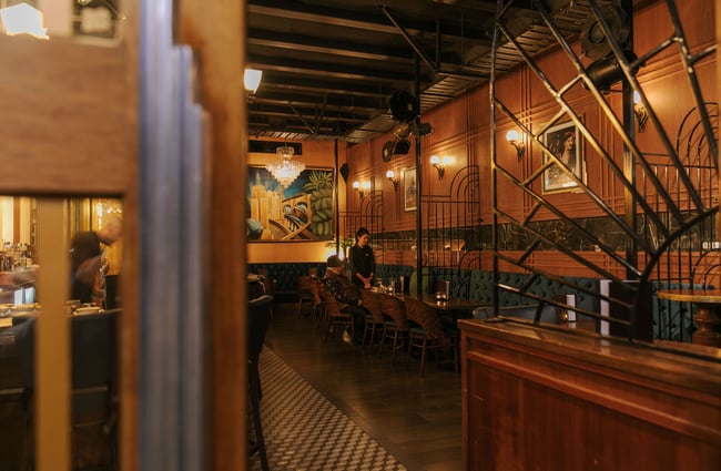 Looking into art deco style bar where a waitress is taking a drinks order from two men surrounded by panelled walls and old school artwork