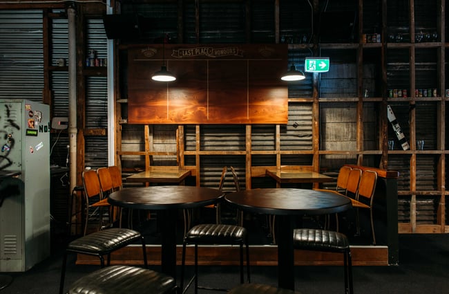 Empty tables and chairs against an exposed wall, lit by two low hanging lights, at Last Place Bar in Hamilton.