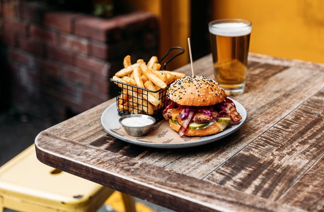 A burger and basket of fries on a plate on a table with a pint of beer behind.