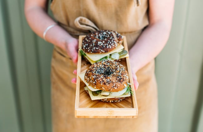 Woman holding a filled bagel on a wooden tray from Little Green Olive, Porirua.