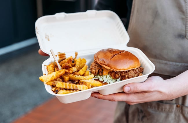 Close-up of a person holding a takeaway box of chips and a fried chicken burger.