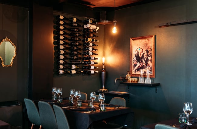 A table and chairs is in the corner by a wine rack mounted on the wall and artwork behind the table at Lustre.