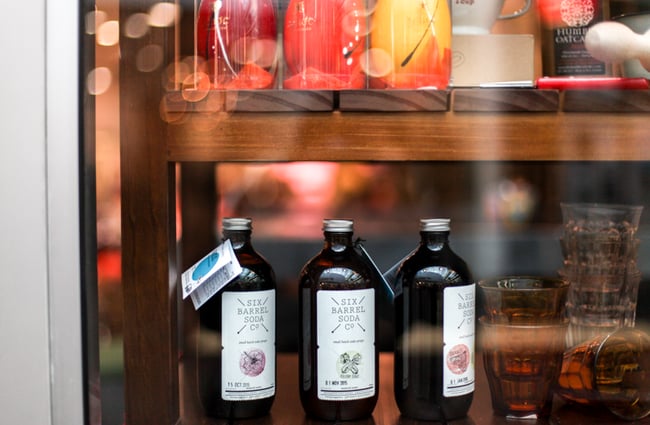 Six Barrel Soda brown glass bottles on wooden shelf.