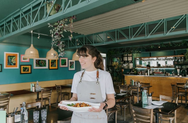 A waitress wearing a grey apron carrying a plate of pasta.
