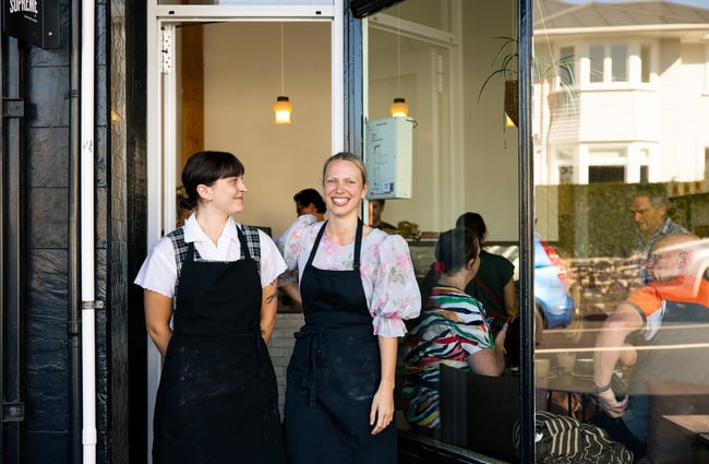 Two women smiling in front of the camera outside a cafe.