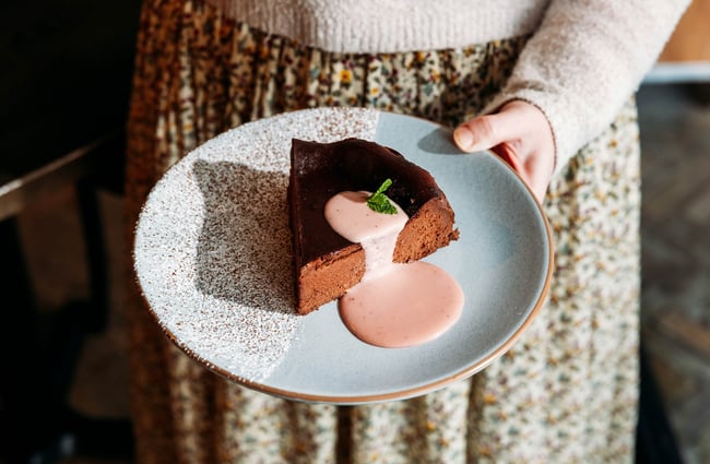 Close up of someone holding out a plate with a slice of chocolate tart on it, with a puddle of pink sauce and two small mint leaves on top.