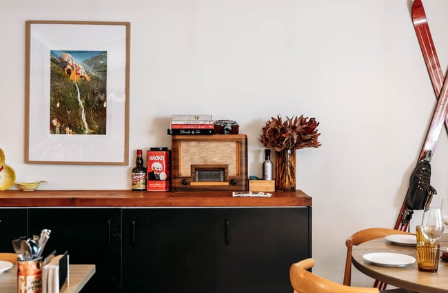 Framed artwork on the wall in the dining area above a wooden shelf with vintage scales, ceramic dishes and big jars of pickles sit in Muttonbird, Wānaka.