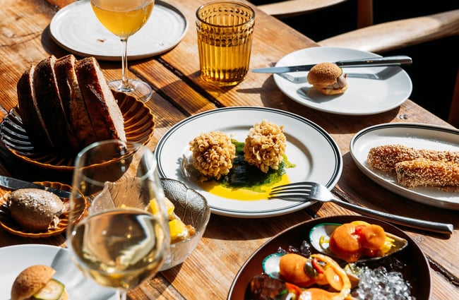 A silver dish of sliced red meat, garlic head halves and a lemon slice and a brown ceramic dish of salad greens sit on the pass at Muttonbird, Wānaka.