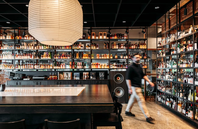 Man walking through the store with bar area in front of him and shelves of alcohol behind him.
