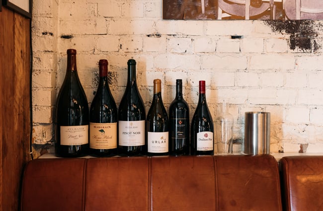 Bottles of wine in a row behind a bench seat in a bar.