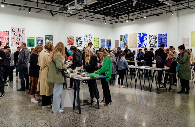 People browsing books and other items on long tables inside a gallery.