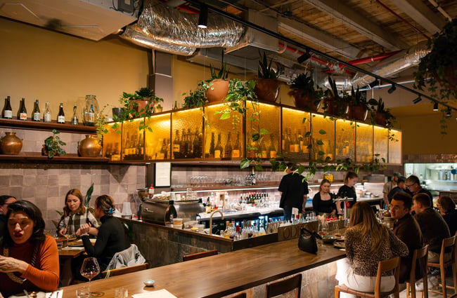 Customers seated at the bar area with bartenders behind them and a view of the open kitchen at Odeon.