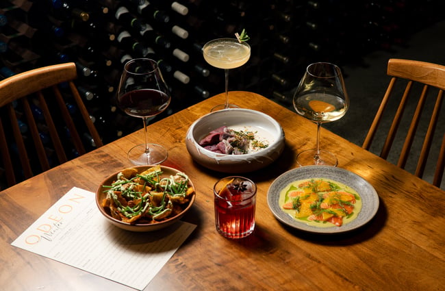 Three different food dishes and cocktails arranged on a table with a menu at Odeon.