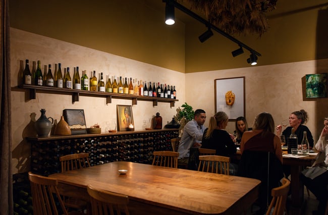 A large group of customers sit at a table above their heads a shelf runs along the wall with wine bottles on it at Odeon.