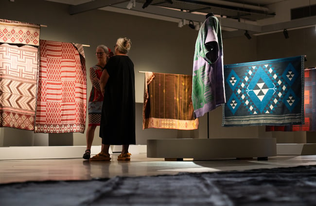 Two women in a dark gallery space surrounded by hanging woven rectangular items.
