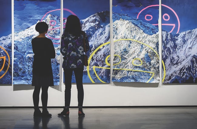 Two women looking at a multi-panel painting of snowy mountain peaks.