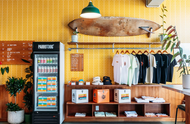 ParrotDog branded apparel hanging on rack under a surfboard next to fridges and a takeaway beer menu