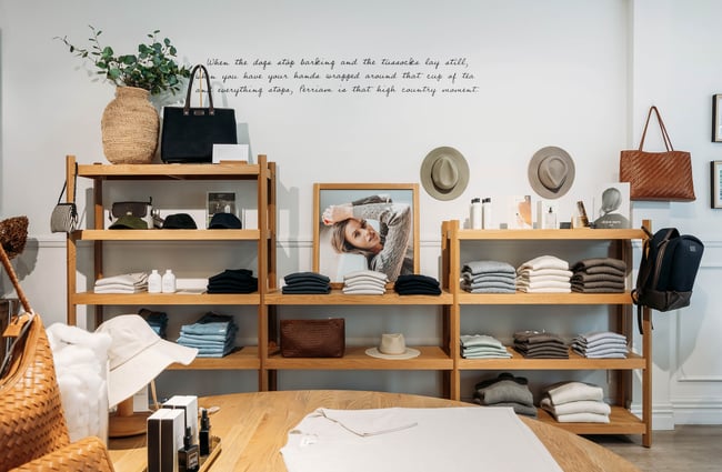Looking across a wooden table to a wooden shelving unit displaying folded clothing, hats and more.