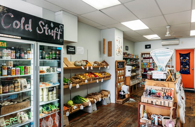 Wider shop view with a row of shelves with products and a fridge in Petal Bee Grocers.