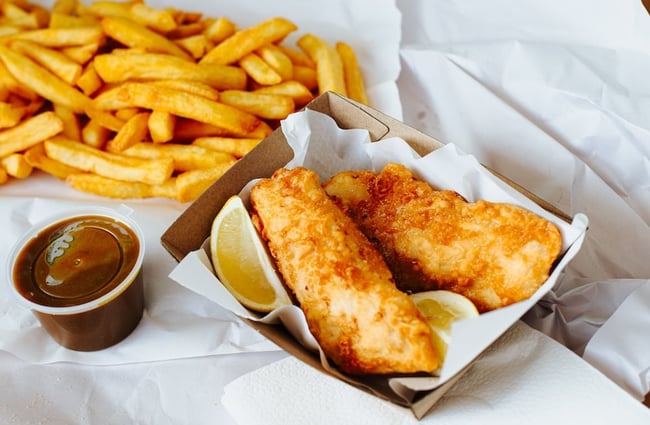 Two pieces of battered fish and lemon slices in a cardboard tray on newsprint paper sheets with chips.
