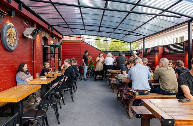 People sitting in the outdoor courtyard at Pomeroy's at wooden tables either side of the space, underneath a curved, clear plastic corrugate roof.