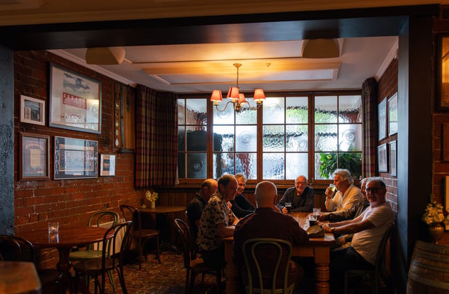 A group of people with pints of beer sit around a large rectangular table inside a nook in Pomeroy's with brick walls on two sides and mottled glass above a timber wainscot on the third side.