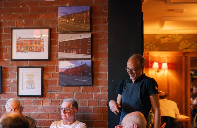 A staff member wearing an apron talks to a table of customers in front of a brick wall with framed photos and paintings of the exterior of Pomeroy's.