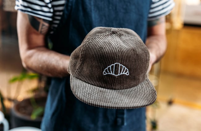 A shop assistant holds a cap with a croissant on it.