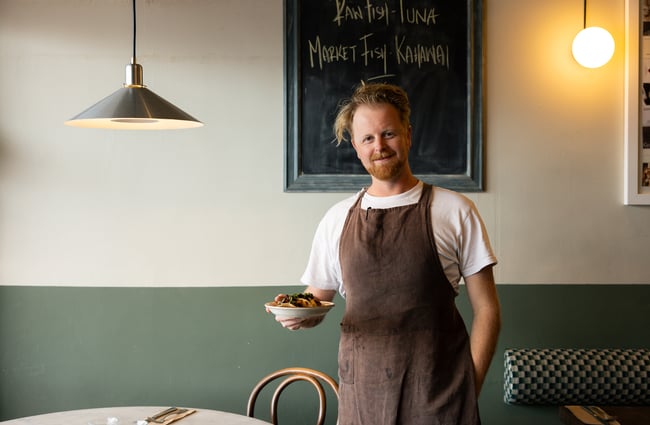 A man wearing an apron and holding a plate of food smiling to camera.
