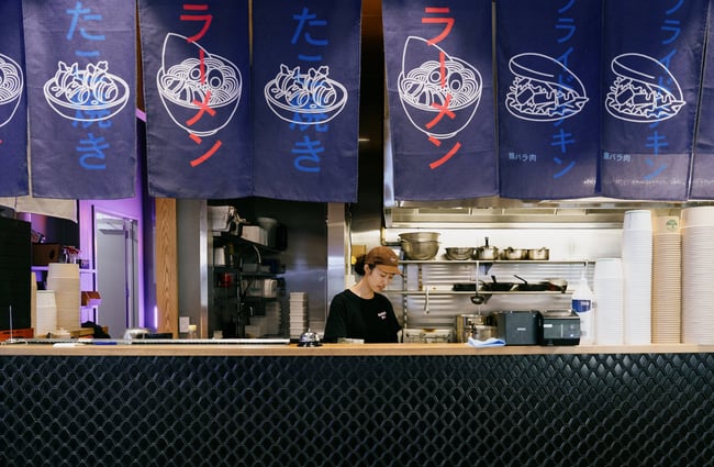 Worker standing behind counter and under Japanese style flags at Ramen Ria in Christchurch