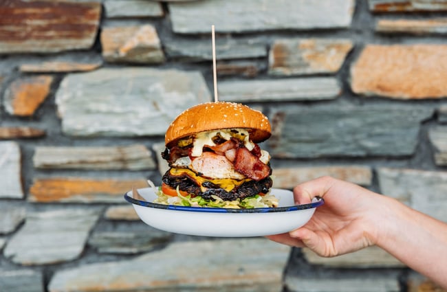 A person holds up an enamel dish with a very tall burger, stacked with bacon, beef, shredded lettuce, tomato, cheese and more, sitting in it.