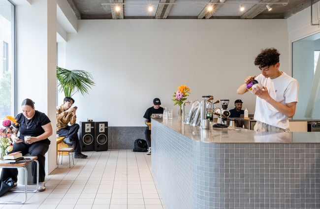 Modern cafe space with barista behind counter making a coffee and customers sitting at tables, in Rook.