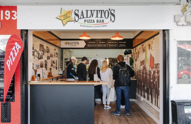 Looking inside from the exterior of the Salvito's Pizza Bar where there is a group of people ordering.