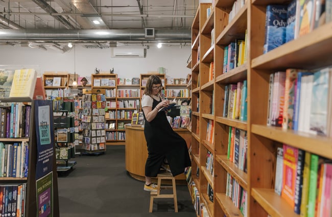 A woman on a stool looks through a book she's taken down from the bookcase.