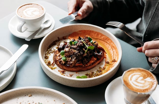 Close up of a milky coffee in a glass and savoury dish in front of a person raising a knife and fork.