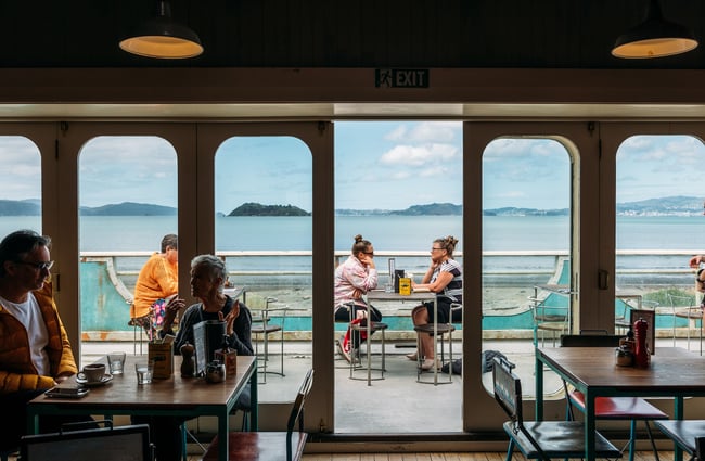 View from inside the first floor Seashore Cabaret cafe out to their deck area where people are dining with a view across the harbour to Wellington city beyond.