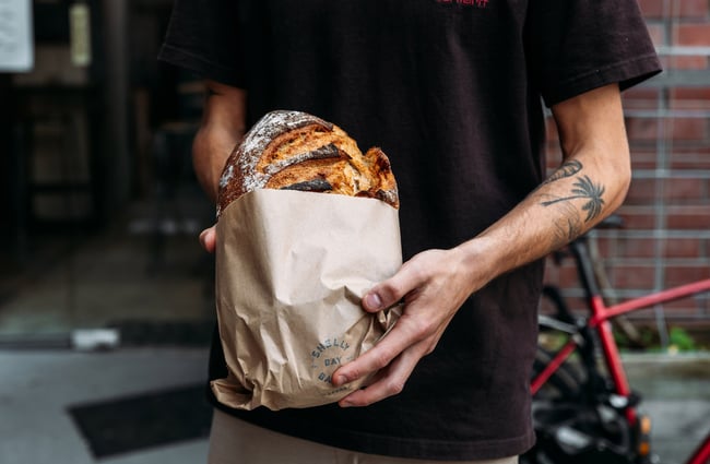 Man with tattooed arm holding a loaf of sourdough bread in brown paper bag