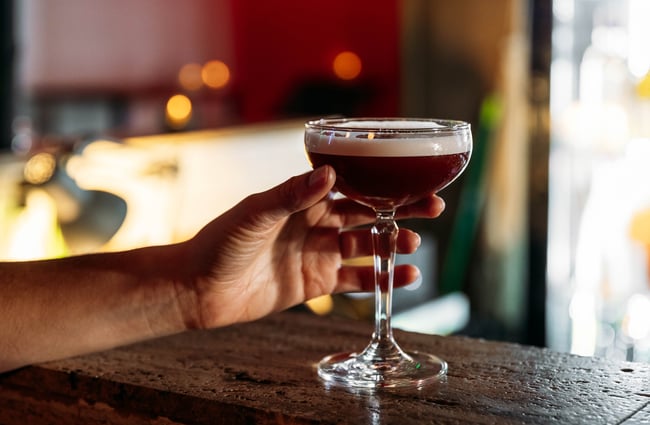 A hand holding an espresso martini on a bar counter.