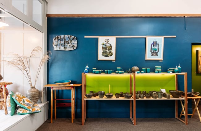 Interior of pottery store with artwork, cushions and a shelf filled with ceramic bowls and mugs glazed in blue and dark tones
