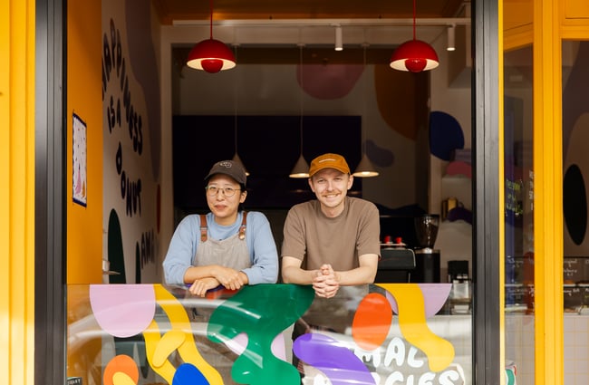 A man and woman looking at the camera from their cafe window.