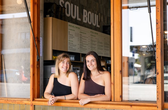 Two women wearing matching tops smiling at the camera through an open window.