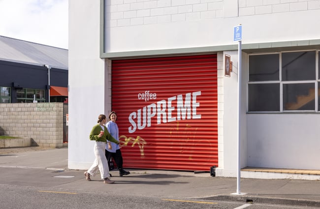Two customers walk past the red roller door with Coffee Supreme signage on it.