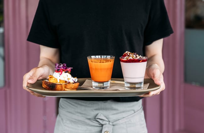 Waiter holding breakfast board with yogurt and fruit, an orange and carrot juice, and a piece of toast with a poached egg and pickles on top